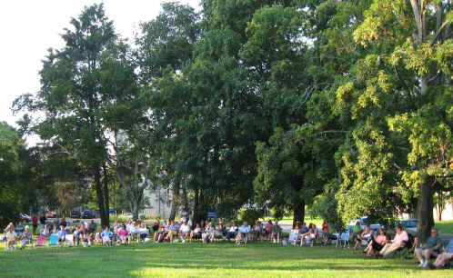 Panaramic view of a hundred neighbors emjoying a summer brass an wind ensemble concert under the trees.